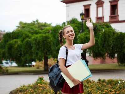 Young beautiful female student smiling, greeting, holding folders outdoors, park background.