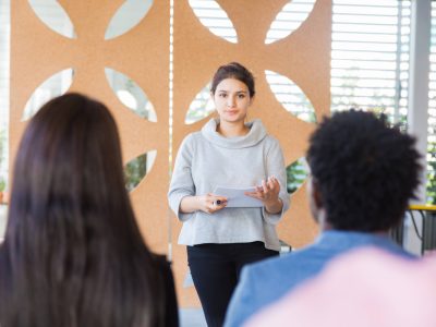 Serious female student presenting project to classmates. Young woman in casual speaking before audience in training room, holding paper with notes. Presentation concept