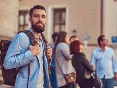 A tourist with a full beard and haircut, wearing casual clothes, holds a backpack and texting on a smartphone, standing on an antique street, during the excursion in Europe.