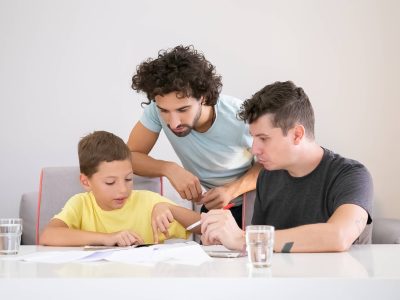 Two dads helping focused boy with school home task, sitting at table with papers, reading textbook together, pointing finger at page. Family and parenthood concept